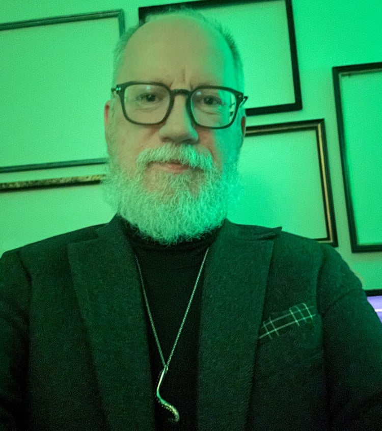 Under an atmospheric green light, David is wearing a dark grey tweed blazer, a black turtleneck, and a silver tentacle pendant.