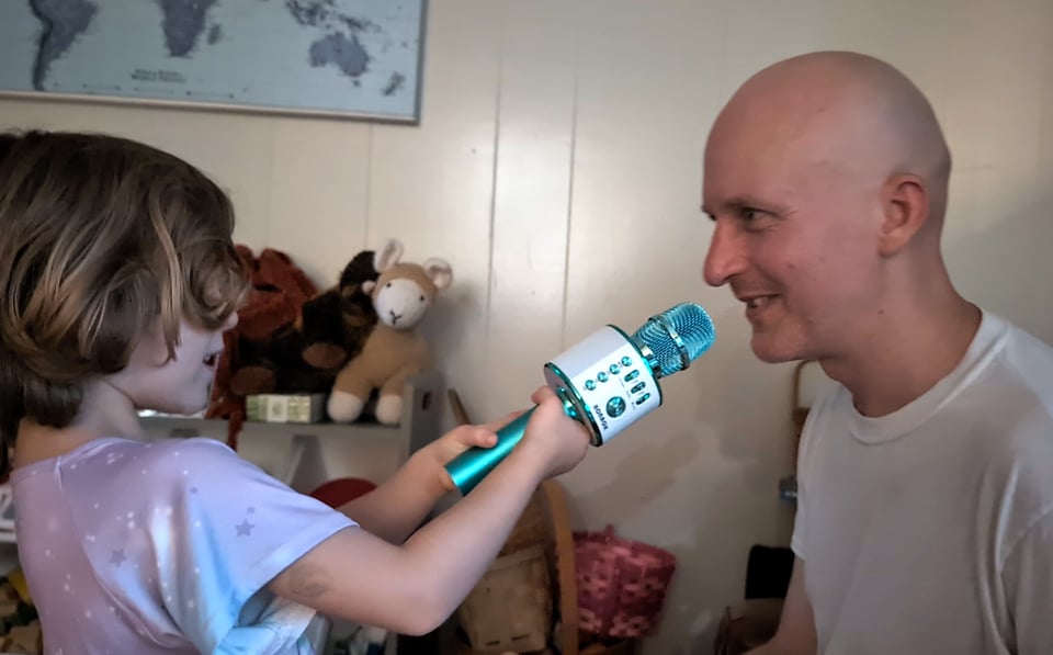 My curly-haired four-year-old holds a sparkly blue toy karaoke microphone up to the mouth of my husband, who is a sweet-natured white man with a shaved head wearing a simple white tee-shirt. Some stuffed animals look on the interview in the background.
