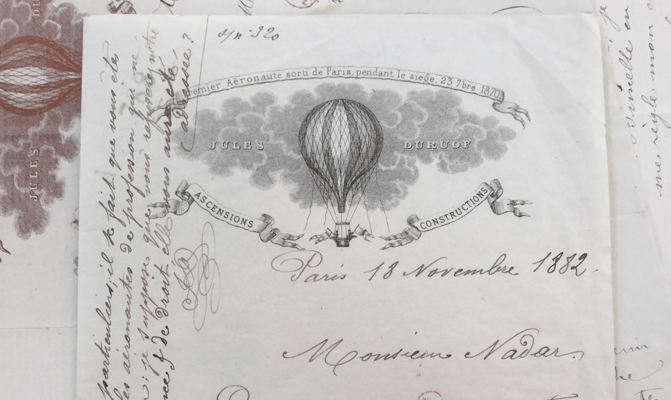 White sheet of paper with printed letterhead in black. The image shows a balloon floating in the clouds with banners advertising the services of Jules Duruof, aeronaut. Handwriting in ink is visible around the image.