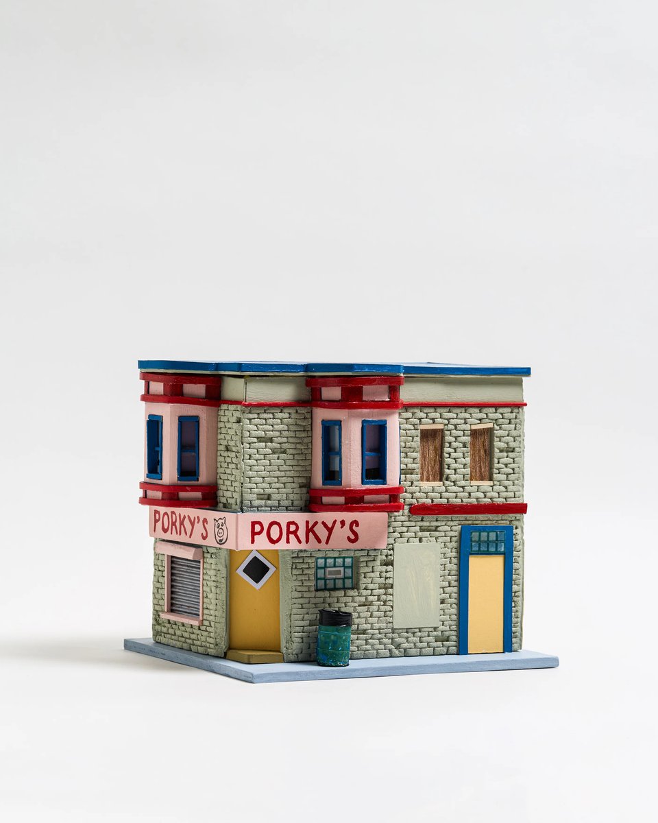 A miniature model of a two-story corner building labeled “Porky’s” on a light pink awning. The awning has a cartoon pig face beside the name. The structure looks like an old-fashioned tenement building from teh 1940s or 1950s, with gray brick walls, vibrant red trim, and blue-framed windows and doors. One yellow door is set at an angle beneath the awning, flanked by a small trash can