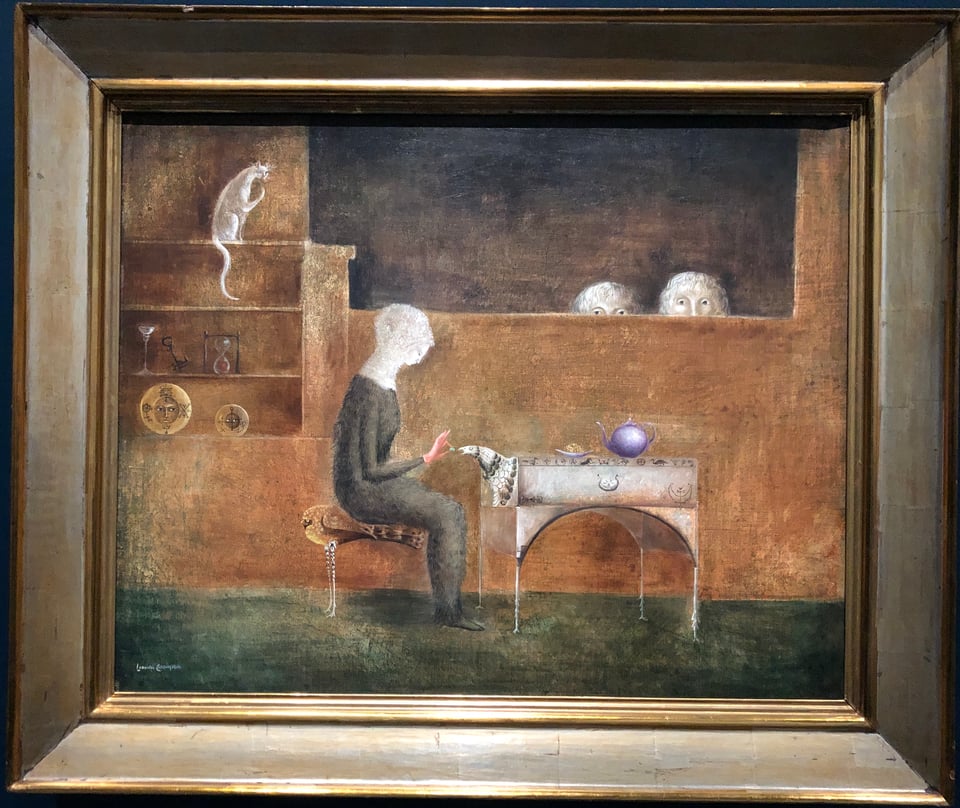 A whimsical painting of a womanly wearing a long, furry dress and furry hat feeding a table with a bird’s head. The figure sits on a bird-shaped stool. Dishes with faces crown down on her from a dresser, while a cat at the top of the dresser languorously considers its options. In the background, the top half of the heads of a pair of children watch the seated figure, all eyes. Most of the painting is in sombre browns, greys, and greens, but there’s a vivid purple teapot on the table.