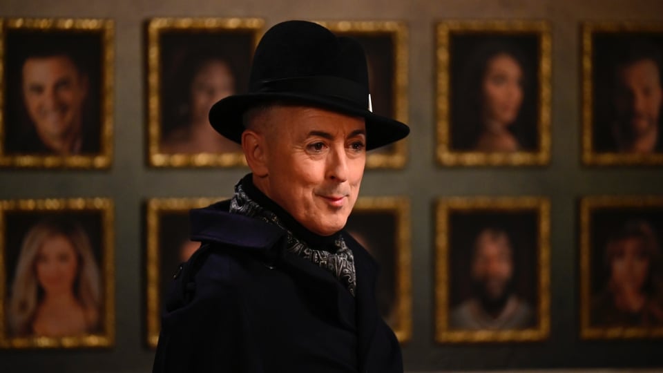 Alan Cumming in a hat, standing in front of a wall of portraits