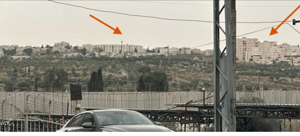 a large gray concrete wall with apartment buildings in the distance, pointed to with orange arrows