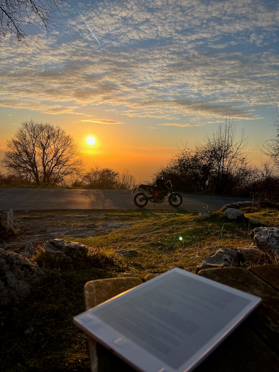An e-ink tablet rests on a wooden table in the foreground, with a motorcycle parked on a roadside in the background along a mountain road against a beautiful sunset with haze and scattered clouds.