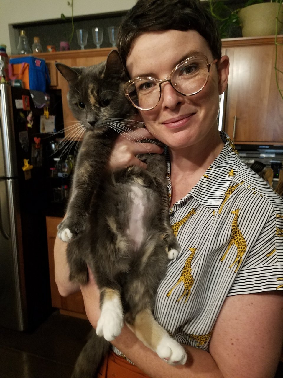 D, a white woman with short dark hair, freckles, and gold glasses, holds a gray dilute tortoiseshell cat with a white belly and hind feet.