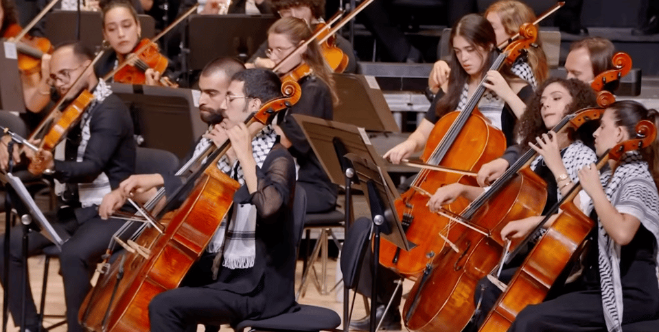 Part of an orchestra, with 10 men and women playing cellos and violins; musicians are all wearing a scarf or other clothing item in the pattern of a Palestinian kuffiyeh