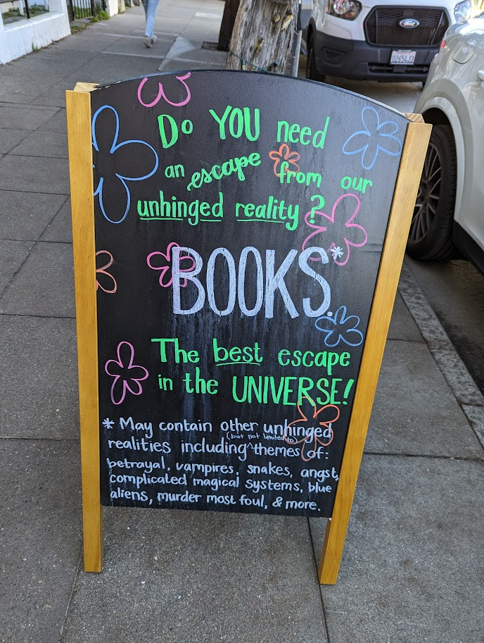 Sandwich board reads: DO YOU need an escape from from our unhinged reality & BOOKS B The best escape in the UNIVERSE! *May contain other unhinged realities including themes (of: betrayal, vampires, snakes, angst, complicated magical systems, blue aliens, murder mast foul, & more.