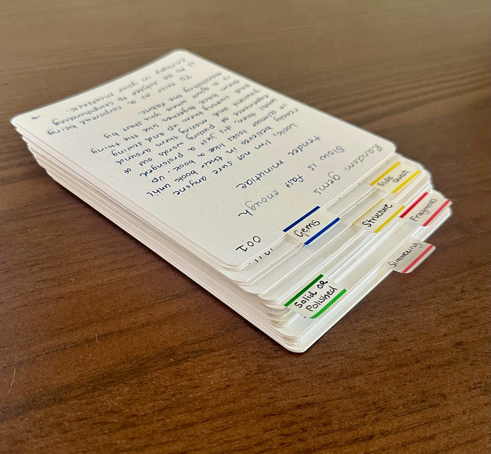 A stack of about 35 index cards with large, tidy writing on the top one facing away from the reader. The cards are staggered to show off the quantity and there are index flag stickers facing the viewer that describe the status of each stack of cards, for example "Simmering", "Fragments", and "Solid OR Polished."