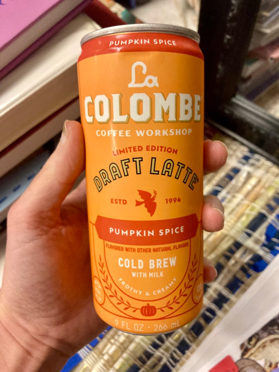 hand holding a canned coffee. the can is orange and the label reads "La Colombe draft latte, pumpkin spice cold brew with milk."
