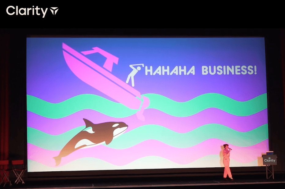 Erika Hall in a pink jumpsuit just like Greta Gerwig's on stage in front of a giant screen with clipart of a golfer on a boat and an orca and the words hahaha business!