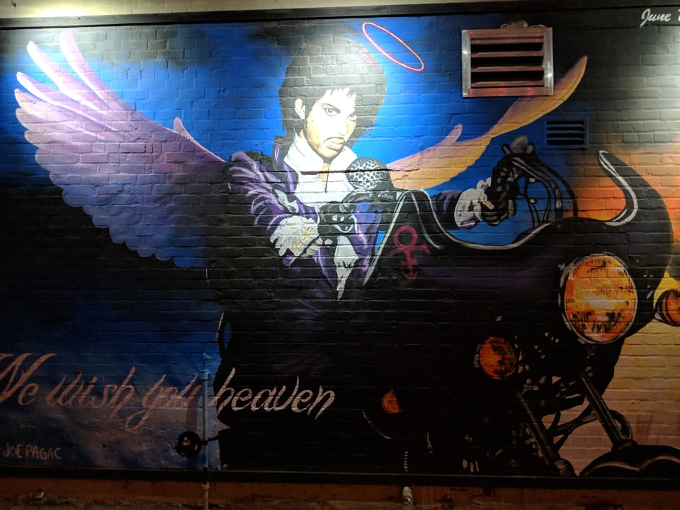 Prince is sitting on his famous motorcycle from Purple Rain, wearing his outfit from that film, with a halo and wings, and it says at the bottom "We wish you heaven".