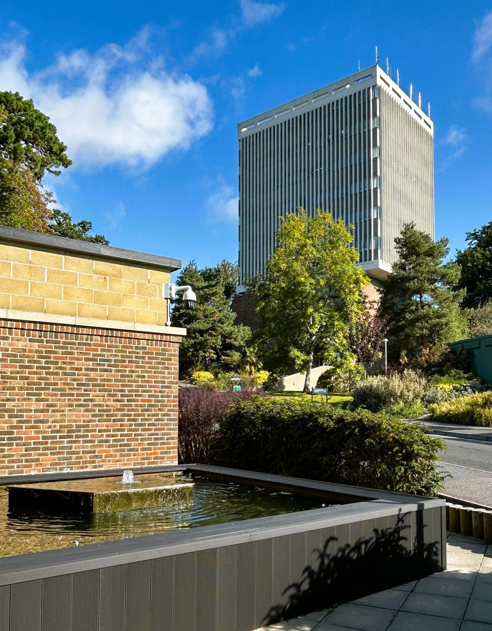 In the foreground is a irregular polygon pool with a small fountain. Across a road, half hidden by mature trees is a pale tower with vertical fins hiding the windows and a white parapet along the top. The lower corner of it can be glimpsed, seemingly floating in the air.