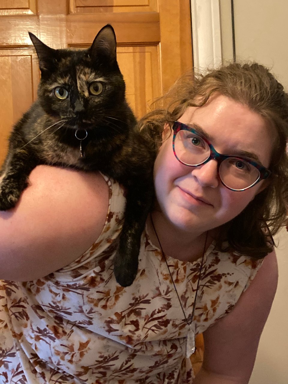 A black and brown tortoiseshell cat with wide yellow eyes is on the shoulder of a nonbinary person in multicolor glasses and a white and brown tunic. The person is bending over slightly to allow the cat to perch more comfortably.