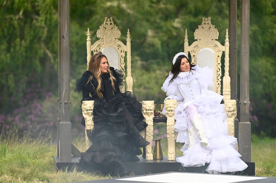 Two people sitting in thrones outside; one is in a black dress and one a white dress