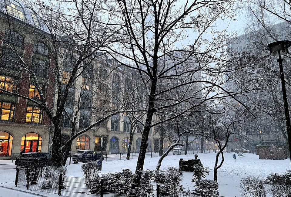 A snow covered small park. There is a stone bench with a carved stone man on it. There are some buildings on the left with yellow lights shining through the windows. It is day-time
