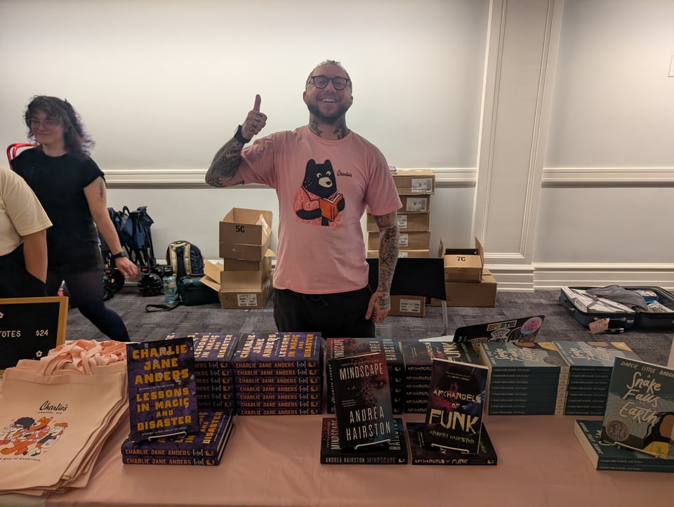 A man with tattoos and a pink t-shirt with a bear reading a book stands with his thumbs up in front of a table covered with books including LESSONS IN MAGIC AND DISASTER, MINDSCAPE, ARCHANGELS OF FUNK AND A SNAKE FALLS TO EARTH.