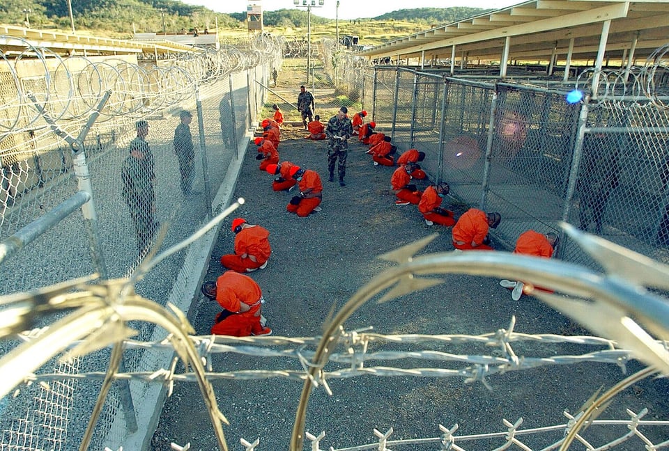 Detainees in orange jumpsuits kneeling on gravel inside a chain link and barbed wire enclosure as members of the military look on.