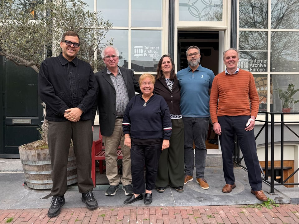 Six people stand in front of a building, all dressed smartly for a meeting and smiling at the camera.