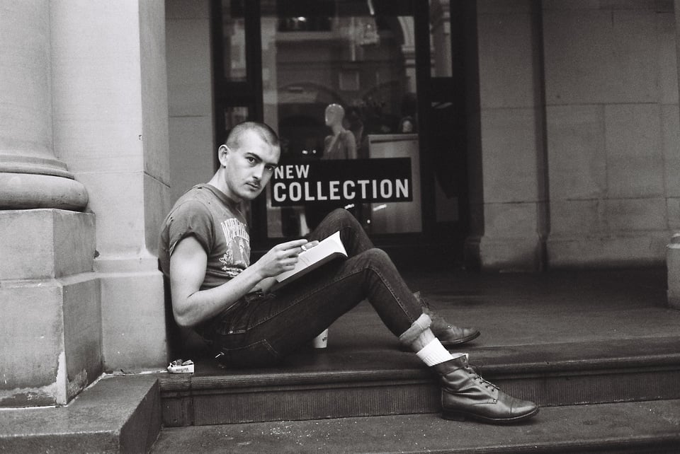 A person with a shaved head sitting on the steps of an old building with a sign in the window that says NEW COLLECTION. They're wearing doc martens and a cool t-shirt and skinny jeans, reading a book and looking at the camera.