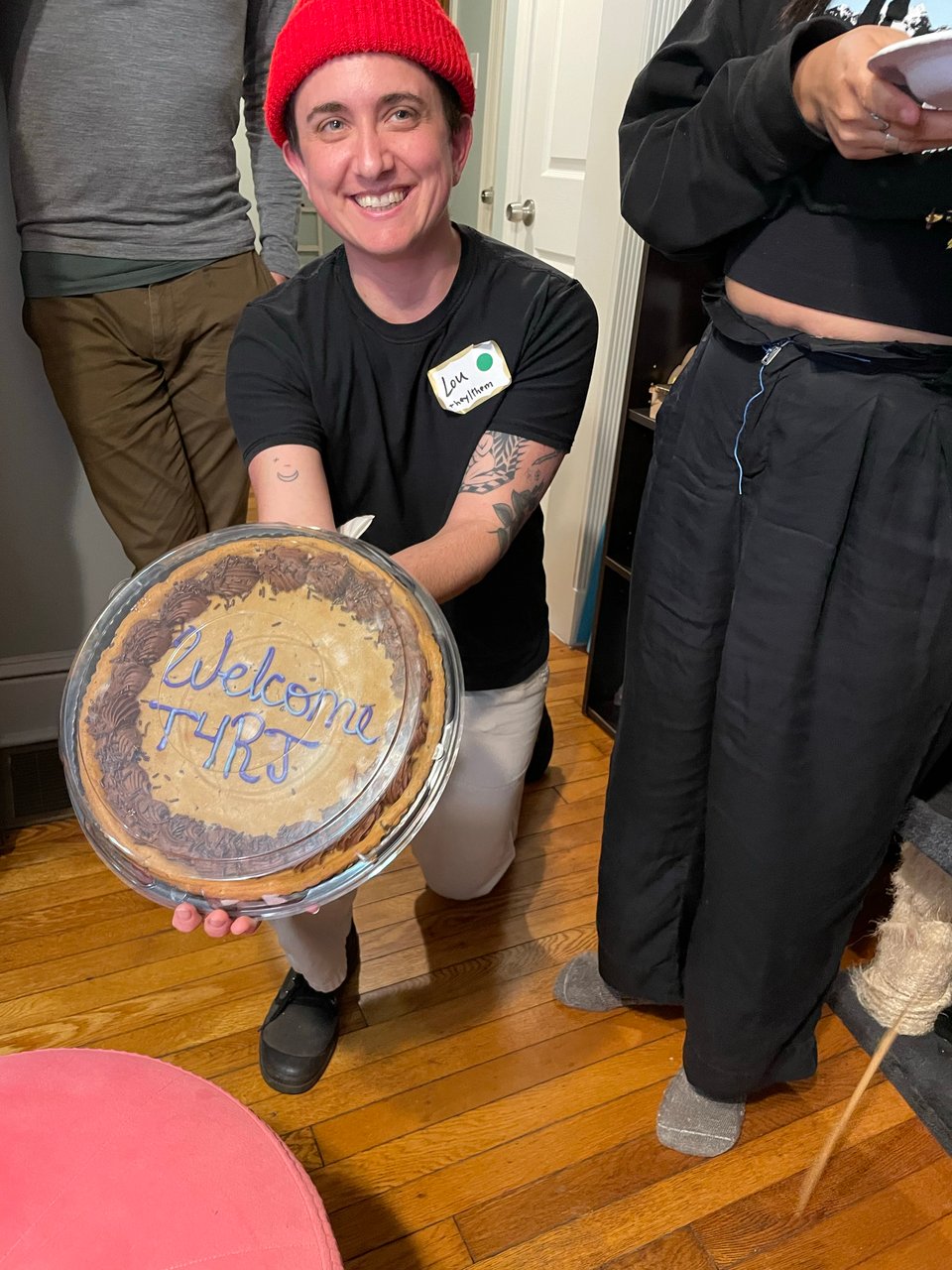 lou on one knee in a red beanie and black tshirt smiling, holding a cookie cake that reads "welcome T4RJ"