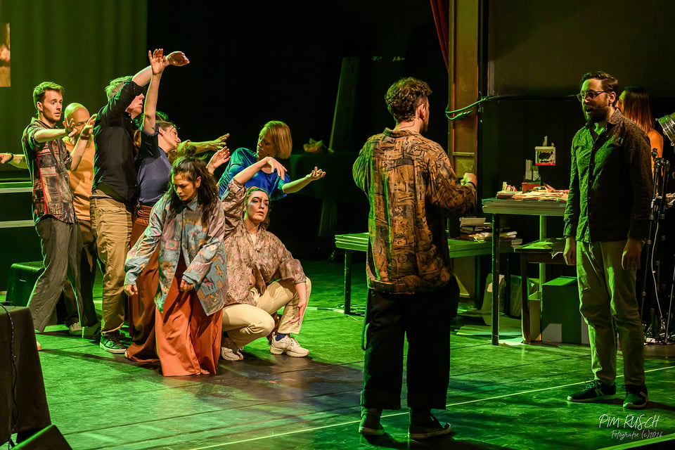 Group of performers on stage, during a Soundpainting performance. Most of them are grouped upstage, dancing, while two stand downstage facing each other.