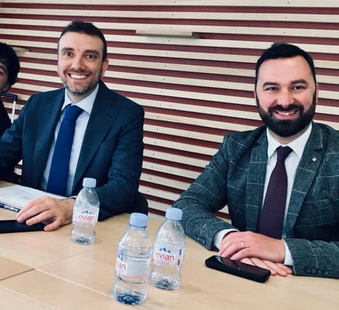 Photo of two smiling men in jackets and ties, seated at a business meeting table with bottled water.