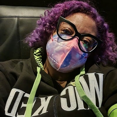 Selfie of Patricia sitting in a movie theatre seat. She is wearing a mask, glasses, and a sweatshirt with the word “Wicked” across the front.