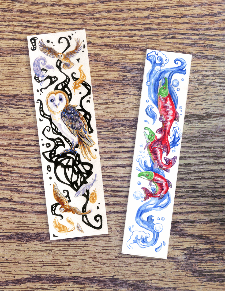 two bookmarks on a wood table surface: the one on the left has yellow and blue barn owls and abstract black ink shapes curling around and behind the owls; the one on the right has sockeye salmon with green heads and red bodies swimming through a curling splash of blue water