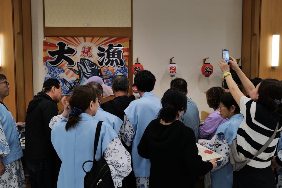 Crowd gathered indoors at a Japanese hotel watching a tuna being butchered, participants in matching blue happi coats with bonito fish motifs facing a bold "great catch" celebration banner, paper matsuri lanterns strung along the wall, a woman raising her phone to capture the moment