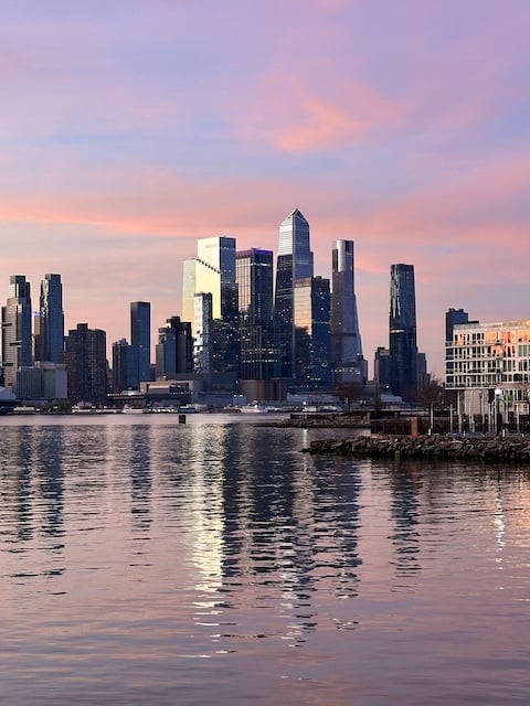 photo of the Hudson Yards area of Manhattan, from across the Hudson River, in early morning sunlight. The buildings are reflected in the river.