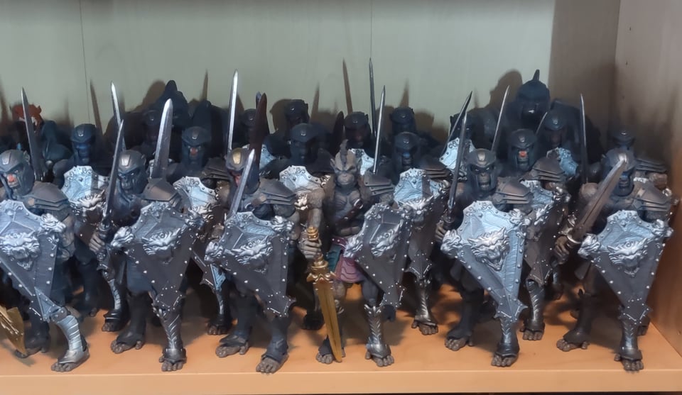A shelf showing, well, nineteen ape-man warrior action figures, all armed with swords and carrying a heavy kite shield. One figure in the front, looking very officer-y, wears no helmet and carries a gold sword. In the back loom two large gorilla figures in armor matching the troops.