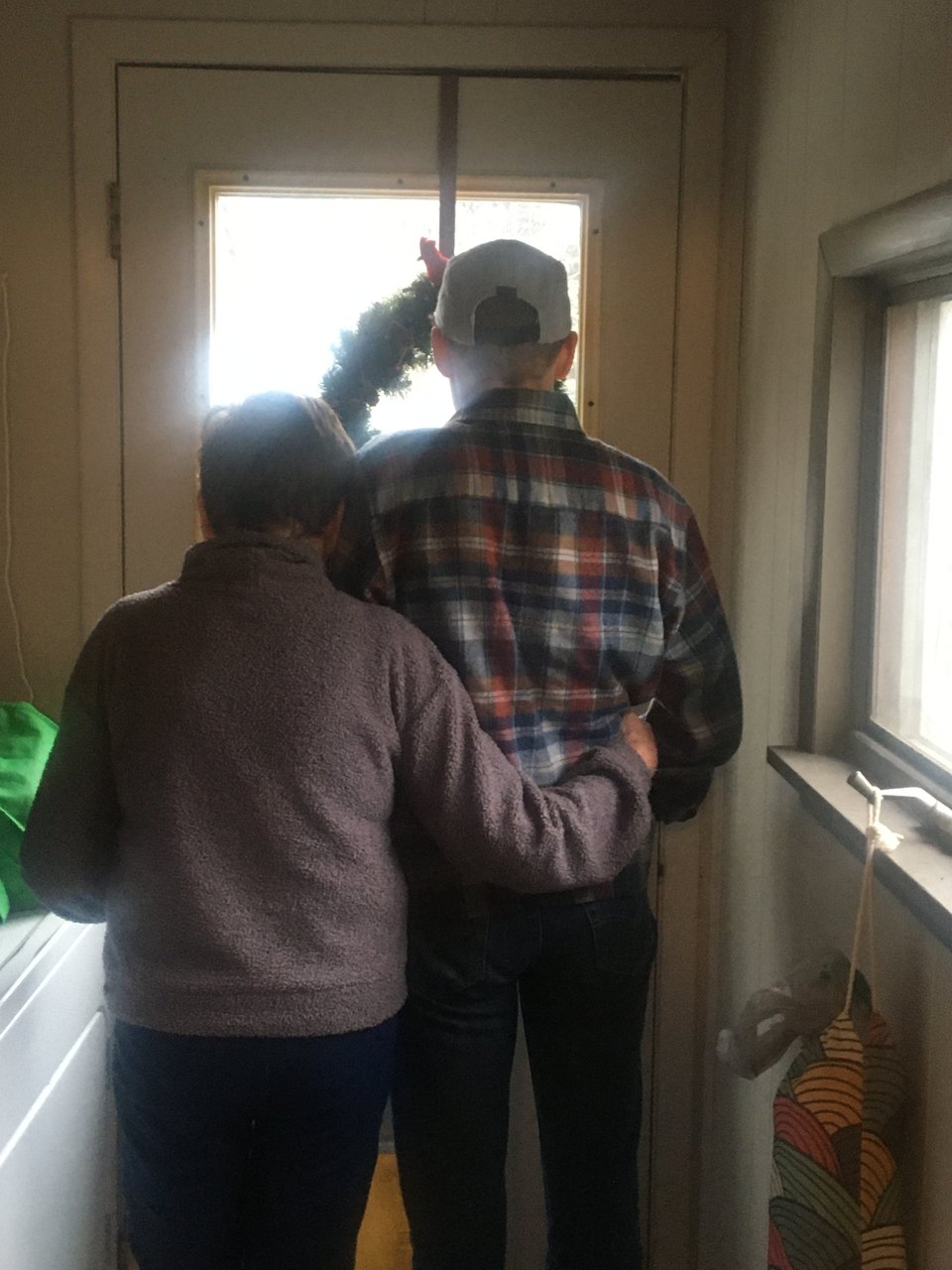 A mom and an adult child stand looking out the backdoor of the house. Mom has an arm around her child's waist