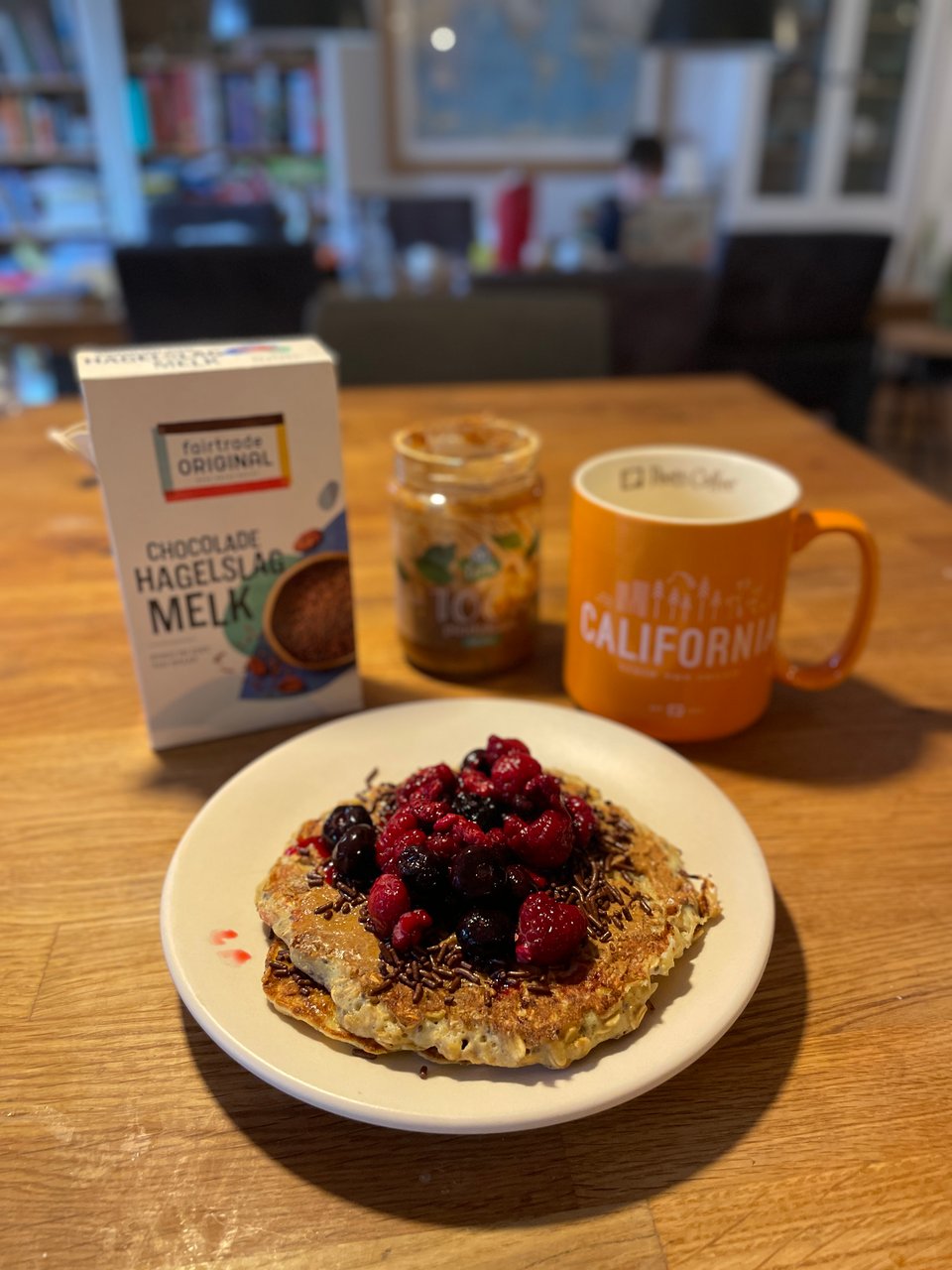 Pancakes are on a plate on a table. They are covered in peanut butter, chocolate sprinkles and berries. Behind the plate is a box of hagelslag (chocolate sprinkles), a jar of peanut butter and a large mug of coffee.
