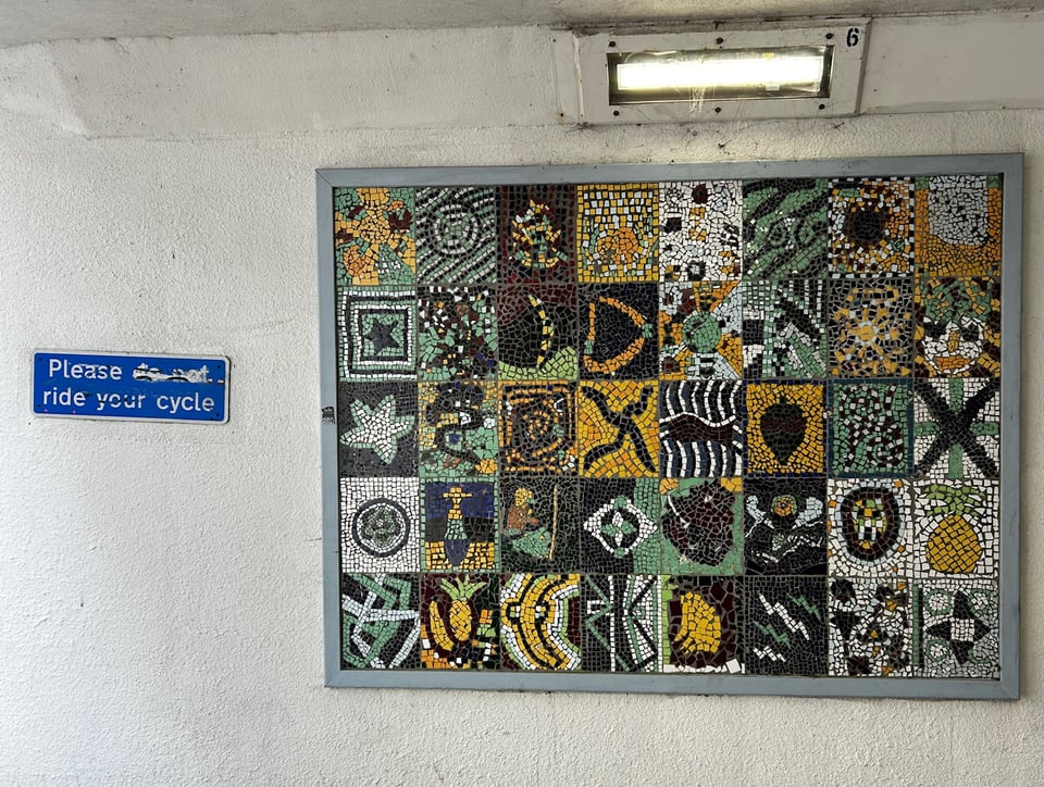 A large mosaic made up of 40 smaller squares all in oches, mint, white and brown. Next to them is a blue sign reading 'Please do not ride your bicycle' but someone has crossed out 'do not' so it reads 'Please ride your cycle'.