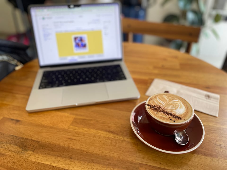 Cappuccino on a table in a brown mug and saucer with a blurred laptop open behind it.