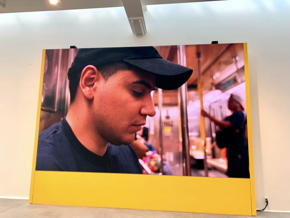 an image of an art gallery installation of a large video showing a person in a baseball cap in a subway train