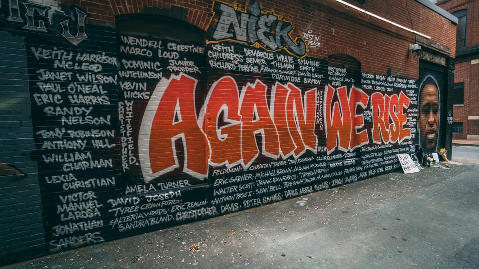 A spraypainted wall, black background, with the names of Black people killed by police, written in white. To the far right of the artwork is a picture of George Floyd. Across the middle, surrounded by the names is the phrase "Again we rise" in orange paint with white drop shadows.
