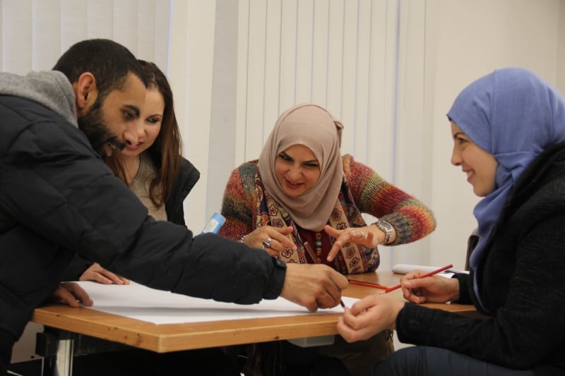 A group of women around a table discuss and draw, with pencils, on a giant white sheet that is being unfurled across a table by a leaning man.