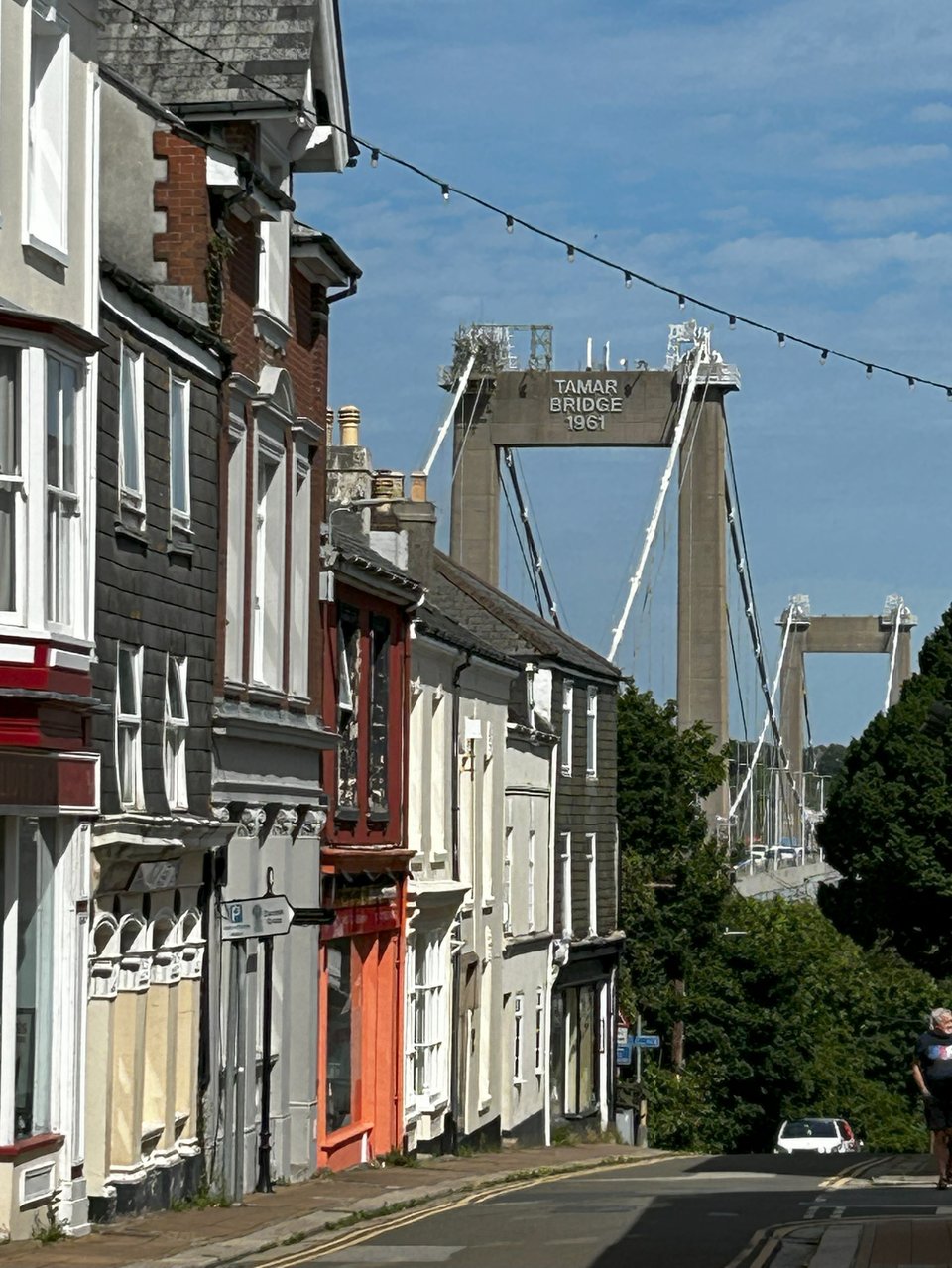 A mishmash terrace of Georgian and Victorian shops and houses descending a hill. Above and beyond them the huge concrete towers of the bridge loom, complete with a sign reading 'Tamar Bridge 1961'.