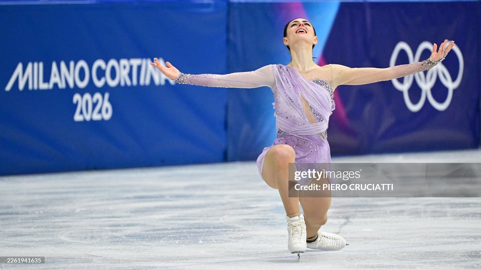 Figure skater in a lavender dress with arms outstretched in a joyous pose on an ice rink, with the Milan Cortina 2026 Olympic logo and branding in the background.