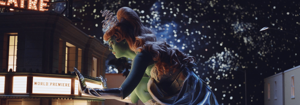 Elle Fanning painted green and dressed as a 50’s style sexy alien peers into a tiny movie theatre. It’s from a particularly lush daydream sequence.