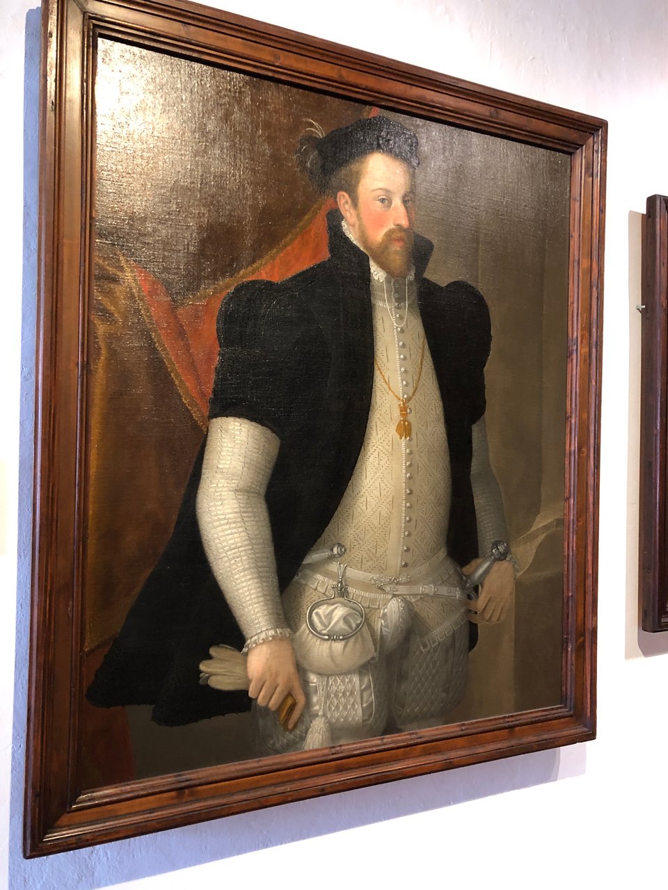 An elegant white man with reddish-brown hair, moustache and beard is in an ivory Renaissance suit and a dark, short-sleeved jacket with a high collar, and a dark floppy hat with a hint or two of a bright red and yellow feather or two. Peeping out from his left side, under the jacket, is the hilt of a sword. On his other side is the handle of (probably) a dagger. Attached to his belt on the right is a small ivory-coloured purse. In his right hand he holds a pair of tan gloves.