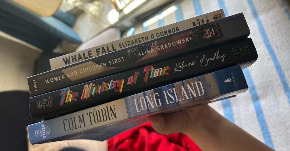 My hand holding up the four novels mentioned below, spine first; in the background, out of focus, a blue and white blanket, a red blanket, and a lamp and blue curtain