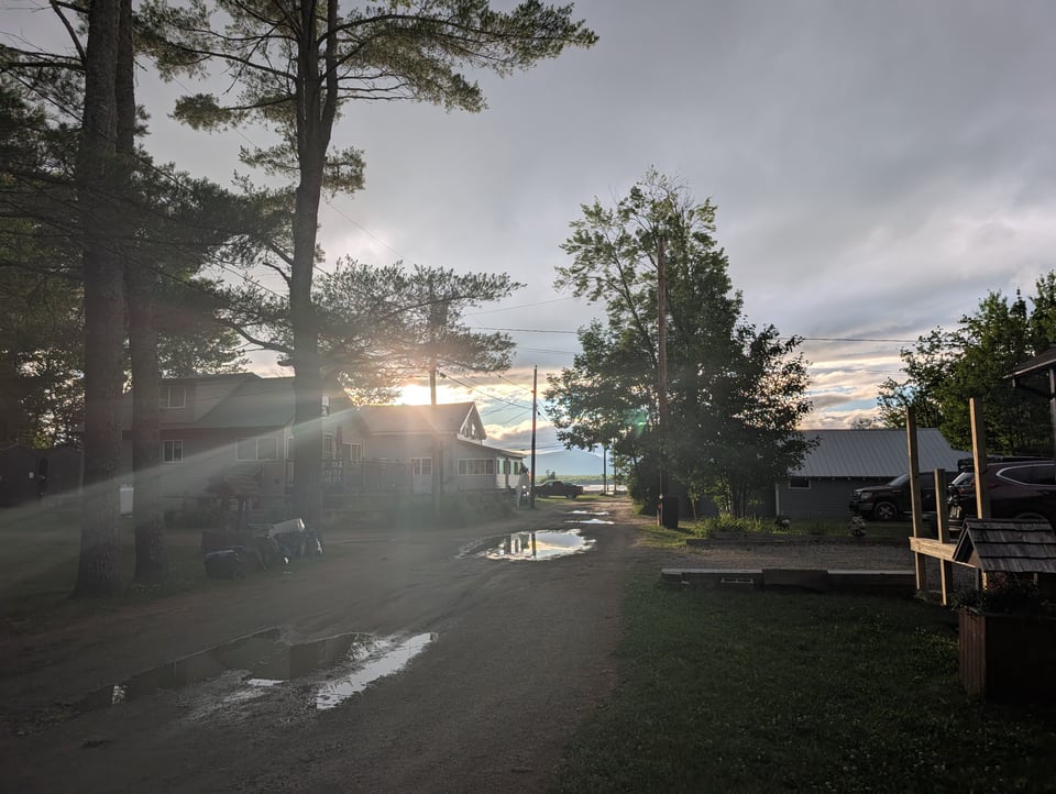 A ray of sun breaking through storm clouds over a tree-lined dirt lane, the sky reflected in its puddles. Small houses on either side stand in silhouette
