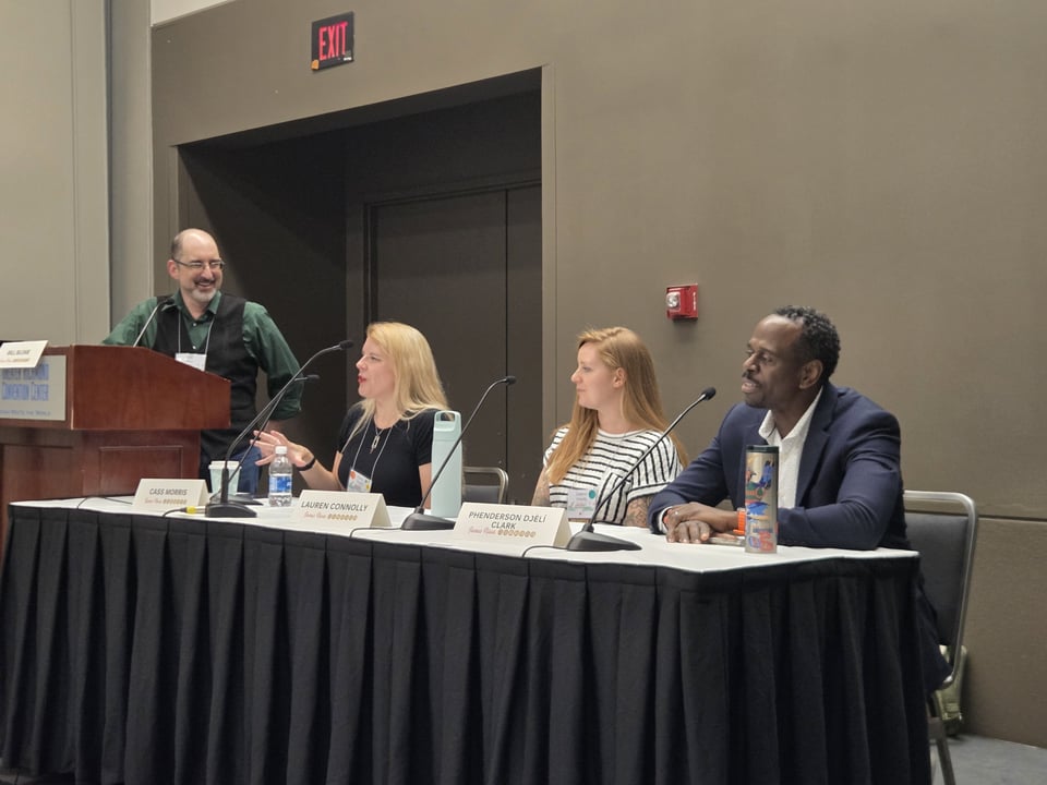 Cass sitting at a table with authors Lauren Connolly and P. Djèlí Clark, with moderator Bill Blume standing at a lectern on her other side. She's in the middle of talking animatedly.