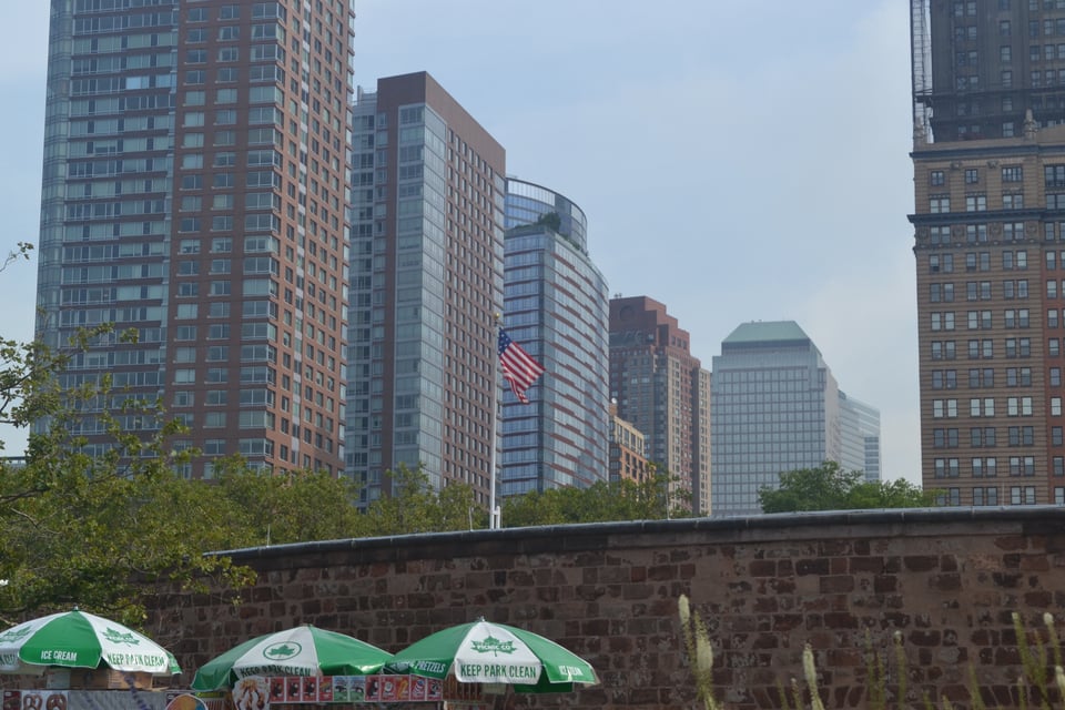 A group of high rise buildings of various designs on the far side of a brick wall. An American flag flies from the top of the wall.