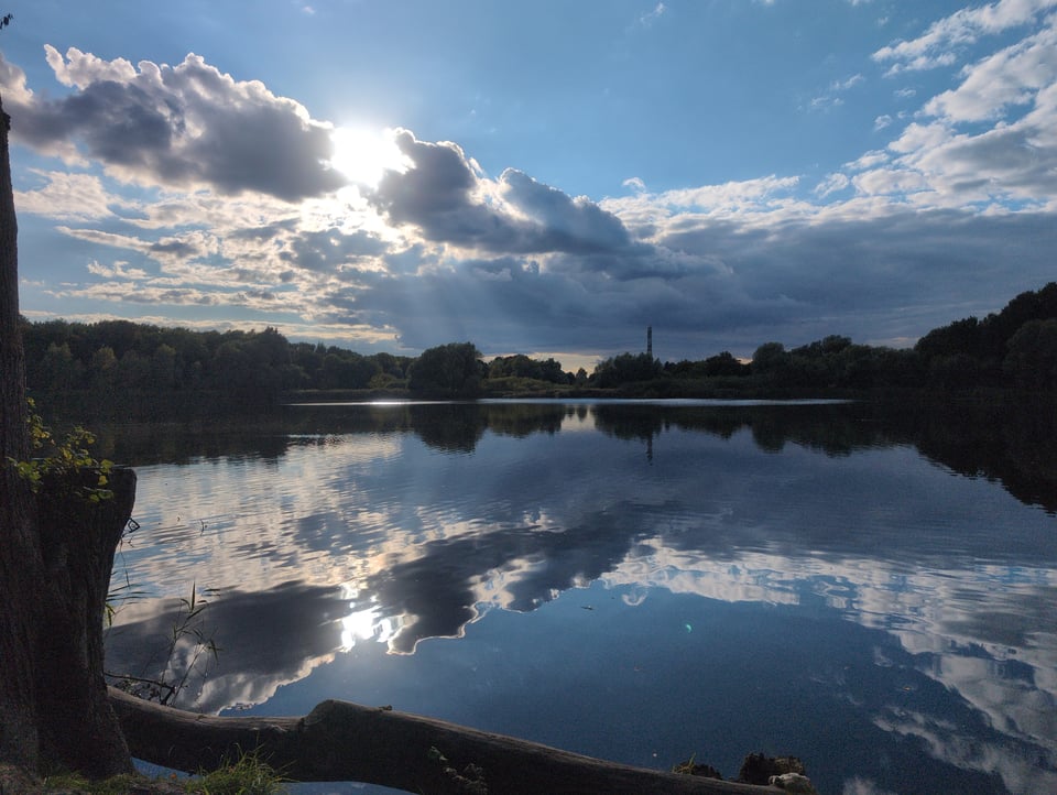 The scene is of a lake. The sky is blue, and clouds curve in the same shape as the lake. The sun glows through the clouds on the left. There are shadows of trees along the far edge of the lake. The clouds, sun, and trees are reflected in the lake.