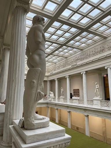 A white plaster cast of a marble sculpture on a balcony overlooking a room below; other statues line the balcony on the opposite side