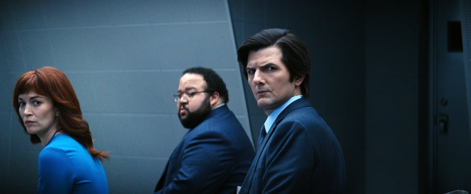 Britt Lower, Zach Cherry and Adam Scott together. in a scene from "Severance," looking toward the camera over their left shoulders with skeptical looks on their faces. The color palette is a pale gray-ish blue of the wall, navy blue in the men's suits and a more vibrant royal blue top hat Lower is wearig.