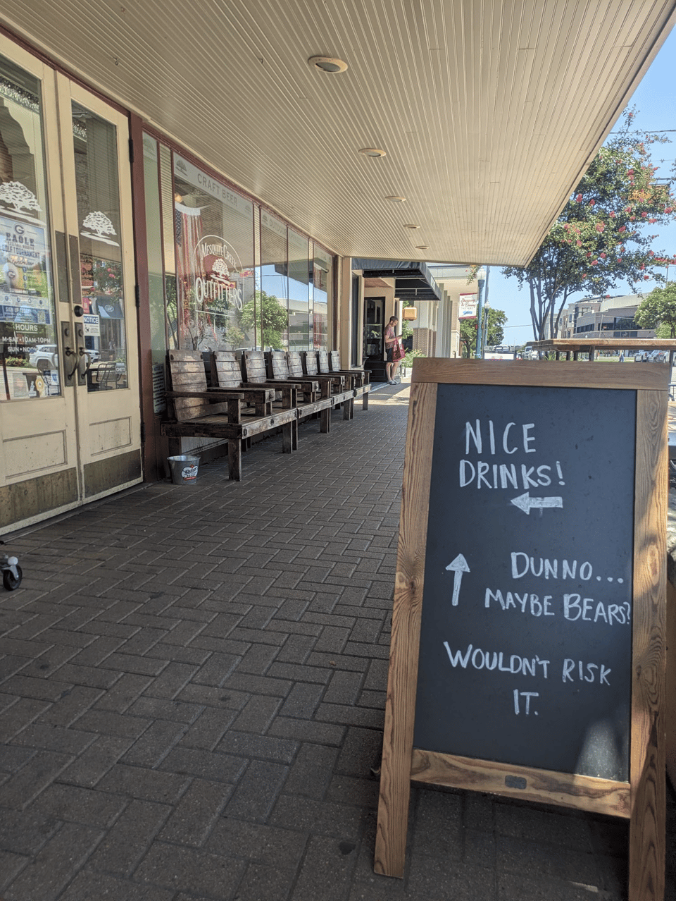 A sidewalk in front of a storefront. The storefront has a row of nice wooden chairs in front of it and big windows with reflection of trees and buildings in them. In the foreground of the image is a sandwich-style chalkboard sign indicating you can turn into the storefront for "Nice Drinks!" or if you go forward there is "Dunno... maybe bears? Wouldn't risk it."